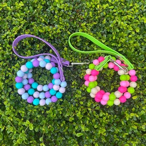 May include: Two beaded dog collars with matching leashes. One collar is purple, blue, and turquoise, and the other is pink, green, and white. Both have silver-tone metal clasps and are set against a green leafy background.