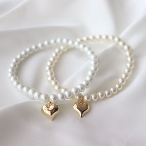 May include: Two pearl bracelets with gold heart charms. The bracelets are white and the hearts are a shiny gold color.