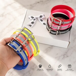 May include: A collection of waterproof dog collars in neon yellow, orange, blue, pink, and black, each featuring a silver metal buckle and a reflective gray strip. The collars are presented with a white gift box and a white ribbon with black paw prints.
