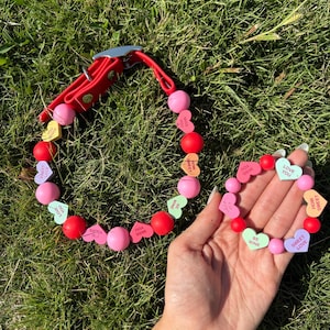 May include: A red dog collar and matching bracelet with pink and red beads. The collar has a silver buckle. Heart-shaped candies with phrases like "Sweet Love" and "Be Mine" are interspersed throughout.