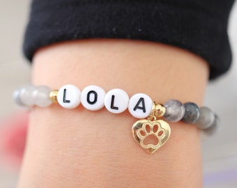 May include: A white beaded bracelet with the name "LOLA" spelled out in black letters. The bracelet has a gold heart charm with a paw print cutout.