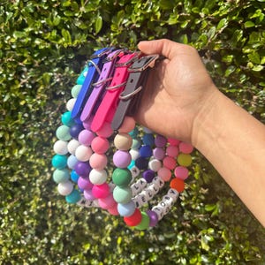 May include: Five colorful silicone beaded dog collars with personalized name tags. The collars are in shades of blue, pink, purple, green, and orange. The names on the tags are "Toby", "Bella", "Layla", "Mia", and "Macy".