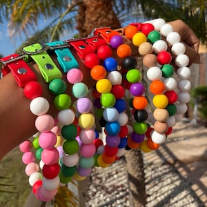 May include: A collection of colorful dog collars with vibrant, round beads in various shades. The collars feature leather straps in red, green, and blue, with silver buckles. The beads are arranged in a gradient of colors, including pink, white, and orange.