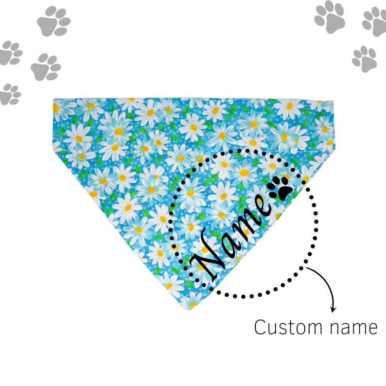 May include: Blue dog bandana with a white daisy print. The bandana has a black dotted circle with the word "Name" and a paw print inside. The text "Custom name" is below the circle.