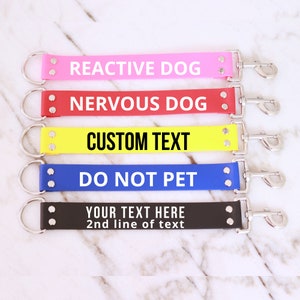 May include: Five different coloured dog leads with metal clips and rings. The leads are pink, red, yellow, blue, and black. The leads have text on them that reads: "Reactive Dog", "Nervous Dog", "Custom Text", "Do Not Pet", and "Your Text Here 2nd line of text".