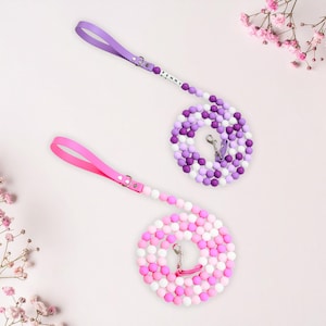 May include: Two beaded dog leashes. One is purple and the other is pink. The purple leash has a matching handle and is made of purple, white, and lavender beads. The pink leash has a matching handle and is made of pink and white beads. The name "PENNY" is spelled out on the purple leash.