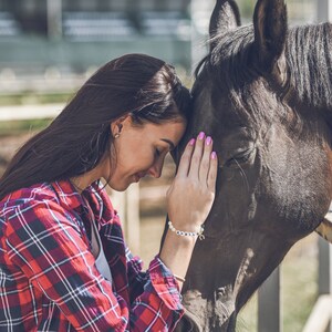 May include: A woman with long brown hair is gently touching the nose of a brown horse. She is wearing a red and white plaid shirt and a bracelet with the word "Apollo" on it.