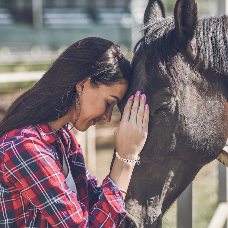 May include: A woman with long brown hair is gently touching the nose of a brown horse. She is wearing a red and white plaid shirt and a bracelet with the word "Apollo" on it.
