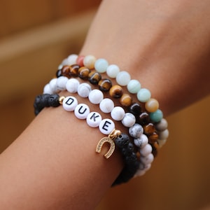 May include: A stack of bracelets with different colored beads, including white, brown, and green. The bottom bracelet has black lava beads and a gold horseshoe charm. The bracelet also has white letter beads spelling out the name "DUKE".