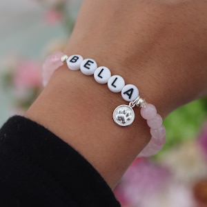 May include: A pink and white beaded bracelet with the name "BELLA" spelled out in white beads and a silver charm with a paw print.