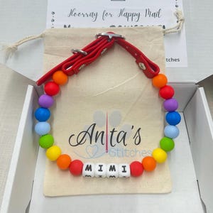 May include: A rainbow-colored dog collar with the name "MIMI" spelled out in white letter beads. The collar has a red leather strap and is packaged in a box with a canvas bag. The bag has the text "Anta's Stitches" printed on it.