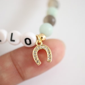 May include: A white beaded bracelet with the word "HELLO" spelled out in black letters. The bracelet has a gold horseshoe charm with clear rhinestones.