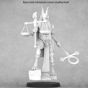 May include: A white, detailed miniature of a jackal-headed figure holding a scale and a staff. The figure is wearing a skirt and a necklace. The miniature is standing on a square base.