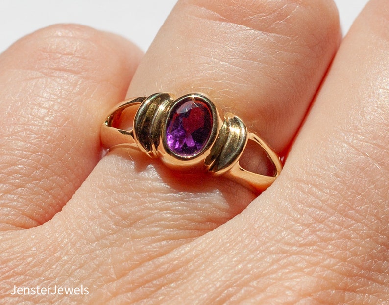 May include: A gold ring with a purple oval gemstone set in a simple, elegant design.
