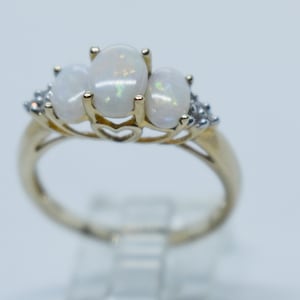 May include: A gold ring featuring three oval opals and small accent stones. The opals have a milky white color with iridescent flashes. The ring's band is a polished gold color, and the setting has a delicate design.