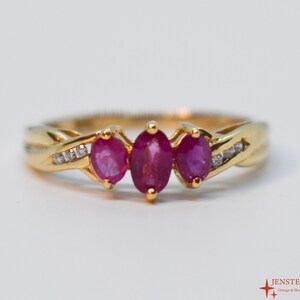 May include: A gold ring featuring three oval-cut rubies and small diamond accents. The ring has a twisted band design and is set against a white background. The ring is a piece of fine jewelry.