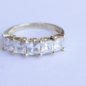 May include: A gold band ring with five clear square-shaped stones set in the band. The ring is engraved with the word "Eternally".