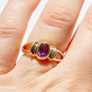 May include: A gold ring with a purple oval gemstone set in a simple, elegant design.