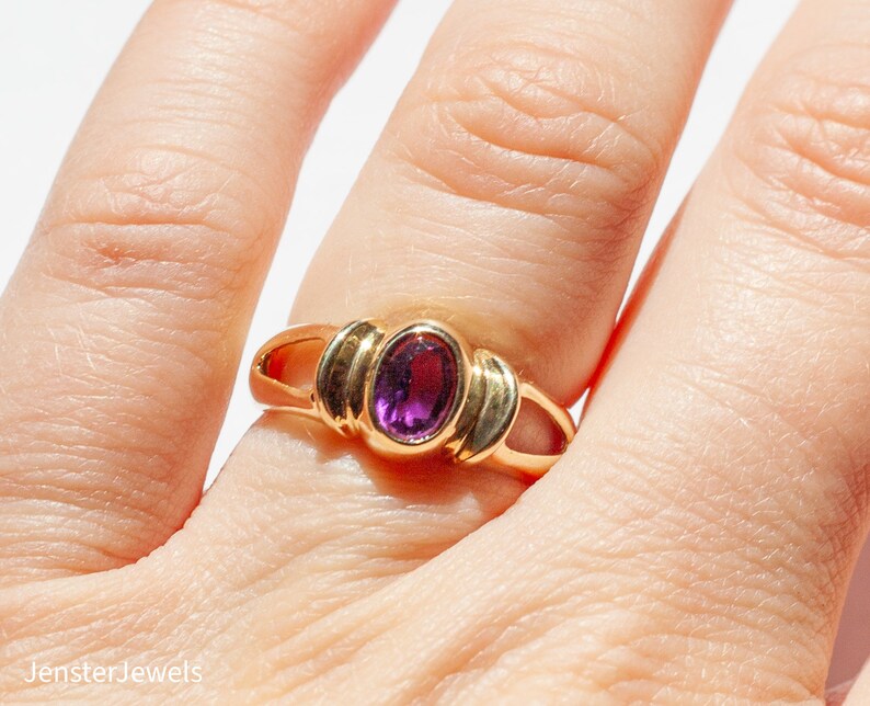 May include: A gold ring with a purple oval gemstone set in a simple, elegant design.