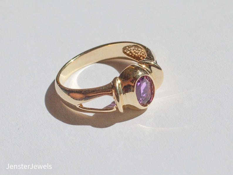 May include: A gold ring with a purple gemstone set in a bezel setting. The ring has a unique design with a twisted band.
