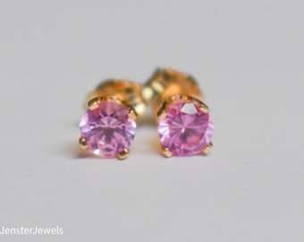 May include: A pair of stud earrings featuring round, light pink gemstones set in gold-colored metal. The earrings are displayed on a white background. The brand name "Jenster Jewels" is visible in the bottom left corner.