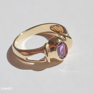 May include: A gold ring with a purple oval gemstone set in a bezel setting. The ring has a simple, elegant design.