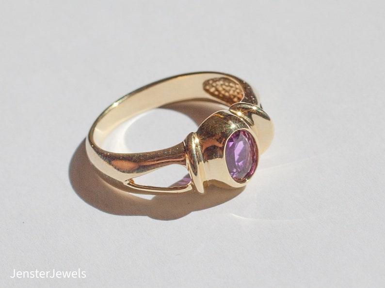 May include: A gold ring with a purple oval gemstone set in a bezel setting. The ring has a simple, elegant design.