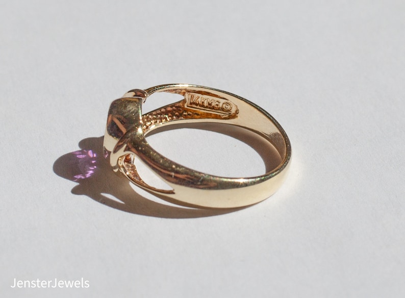 May include: A gold ring with a purple gemstone set in a knot design. The ring is marked "14K" on the inside of the band.