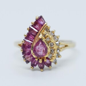 May include: A gold ring featuring a teardrop-shaped cluster of pink gemstones and surrounding smaller white stones. The ring's design includes a fan-like arrangement of pink stones and a central, pear-shaped pink stone.