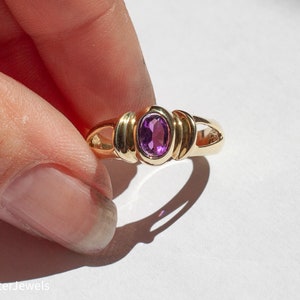 May include: A gold ring with a purple oval gemstone set in the center. The ring has a simple, elegant design.