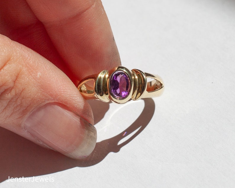 May include: A gold ring with a purple oval gemstone set in the center. The ring has a simple, elegant design.