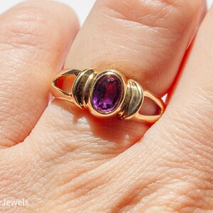 May include: A gold ring with a purple oval gemstone set in a bezel setting. The ring has a simple design with a band that is slightly wider than the setting.