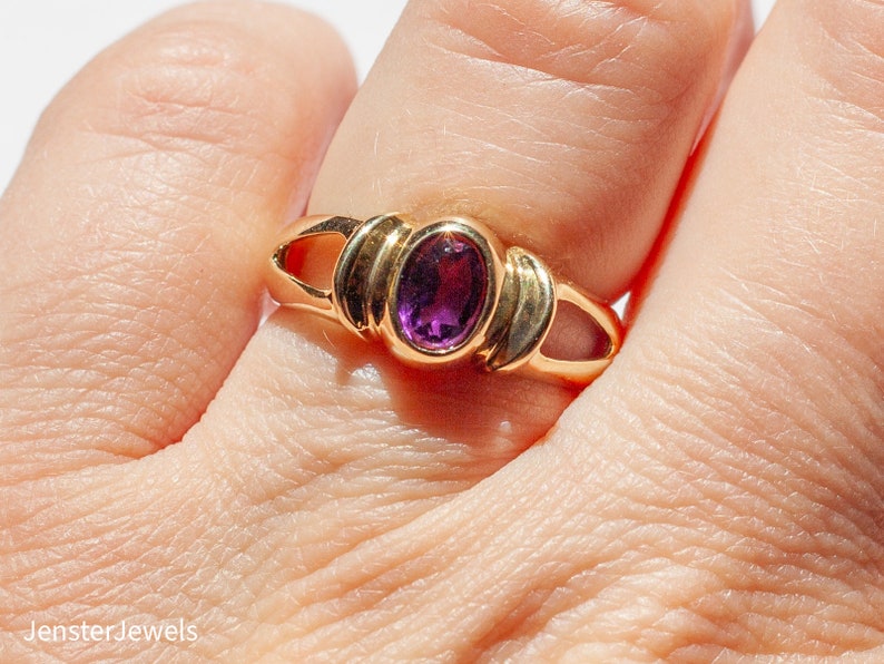 May include: A gold ring with a purple oval gemstone set in a bezel setting. The ring has a simple design with a band that is slightly wider than the setting.