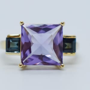 May include: A gold ring featuring a large, square-cut, light purple gemstone as the centerpiece. Two smaller, rectangular, dark blue gemstones flank the main stone. The ring has a classic, elegant design.