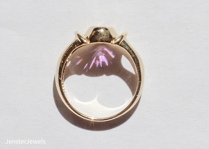 May include: A gold ring with a large, clear gemstone that has a purple hue. The ring is set in a bezel setting and has a simple band.