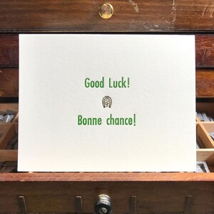 Pode incluir: Um cartão branco com texto verde que diz "Good Luck!" e "Bonne chance!" com um gráfico de ferradura. O cartão é exibido em uma gaveta de madeira.