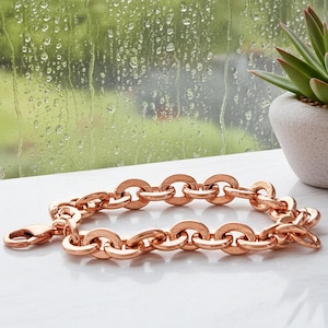 May include: Rose gold chain link bracelet with a lobster clasp. The bracelet is made of connected oval links. The bracelet is displayed on a white surface with a blurred background of a window and a potted succulent.