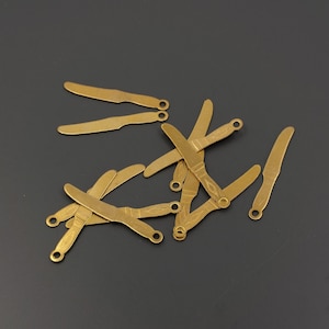 May include: A group of ten gold-colored metal knife charms, each with a slightly different design. The charms are arranged in a pile on a dark gray surface.