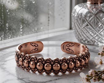 May include: A copper cuff bracelet featuring a repeating pattern of raised circular beads and scalloped edges. The interior of the open-ended band is engraved with small lightning bolt symbols and geometric markings.