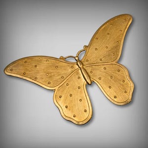 Puede incluir: Un objeto decorativo en forma de mariposa de color dorado. La mariposa tiene alas texturizadas con pequeños detalles circulares espaciados uniformemente. El cuerpo y las antenas también son dorados. El objeto probablemente está hecho de metal y se muestra sobre un fondo gris degradado.