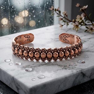 May include: A copper cuff bracelet featuring a central row of polished spherical beads bordered by scalloped edges. The interior ends of the open band are engraved with lightning bolt symbols and decorative geometric patterns.