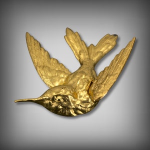Peut inclure: Une sculpture d'oiseau-mouche en métal doré aux ailes déployées. La sculpture est détaillée avec des plumes texturées et un bec délicat.