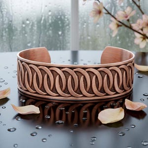 May include: A copper cuff bracelet featuring a repeating pattern of interlocking oval shapes with a textured background. The metal has a brushed finish and a wide, open-ended band design that curves into a circular shape.