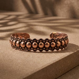 May include: A copper cuff bracelet featuring a central row of raised, polished spheres bordered by scalloped edges. The metal surface displays a warm, metallic reddish-brown tone with intricate stamped markings visible along the inner band.