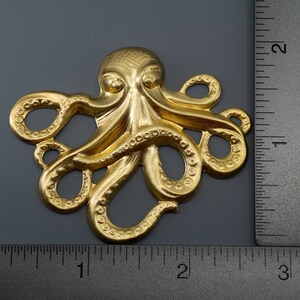 K&ouml;nnte beinhalten: Ein goldfarbener Metalloktopus mit strukturierten Details. Der Oktopus ist etwa 5 cm breit und wird neben einem Lineal zur Gr&ouml;&szlig;enbestimmung gezeigt.