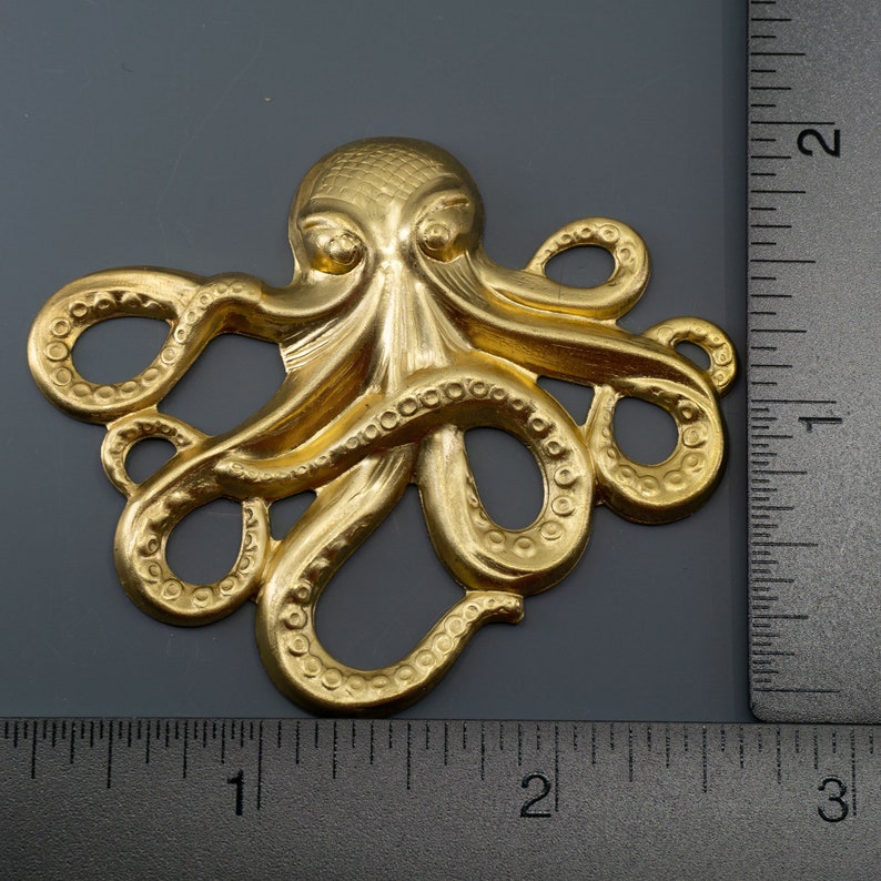 K&ouml;nnte beinhalten: Ein goldfarbener Metalloktopus mit strukturierten Details. Der Oktopus ist etwa 5 cm breit und wird neben einem Lineal zur Gr&ouml;&szlig;enbestimmung gezeigt.