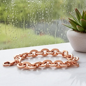 May include: A rose gold chain bracelet featuring thick, rounded oval links and a secure lobster claw clasp. The metal has a polished, reflective finish that catches the light, creating a warm metallic tone across the interconnected circular links.