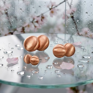 May include: Two pairs of round, brushed copper-toned stud earrings. The larger pair features two spherical shapes, while the smaller pair consists of two smaller spheres. Each earring has a textured, matte metallic finish with a warm, reddish-brown hue.