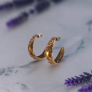 May include: A pair of gold-toned hoop earrings featuring an embossed floral pattern across the exterior surface. The hoops have a C-shaped design with a polished finish and clear plastic earring backs attached to the posts for secure wear.