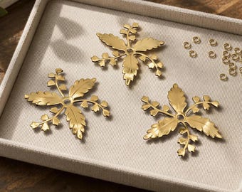 Puede incluir: Tres fornituras de joyería de metal dorado con forma de hoja y un orificio central, acompañadas de una pequeña colección de anillas de salto doradas. Los componentes presentan un diseño botánico detallado con bordes serrados.